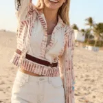 Modella sorridente sulla spiaggia, indossa una giacca a righe e pantaloni corti chiari, mentre alza il braccio in segno di felicità. Sfondo con palme e cielo sereno.