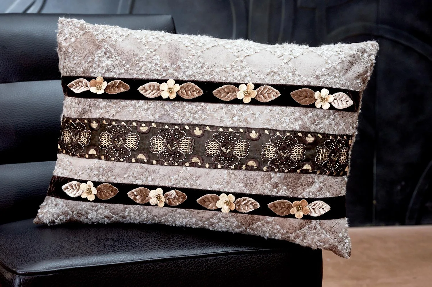 Cuscino decorativo con tessuto morbido e dettagli floreali, posizionato su un divano nero. Design elegante con strisce di velluto e ricami che aggiungono un tocco di stile all'arredamento.