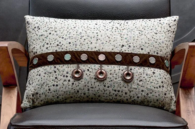 Cuscino decorativo con design elegante su una sedia in pelle nera, caratterizzato da un tessuto grigio con dettagli glitterati e una fascia marrone con bottoni metallici. Perfetto per aggiungere stile e comfort agli interni.