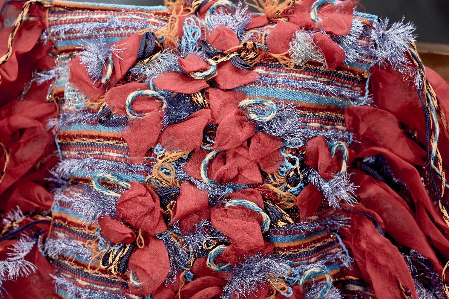 Dettaglio di un tessuto artistico con fiori in stoffa rossa e filamenti blu e metallici, evidenziando la complessità e la bellezza del design.