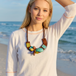 Modella con un collo di perle colorate e una maglietta bianca, in piedi sulla spiaggia con il mare sullo sfondo. La luce del sole illumina la scena, creando un'atmosfera estiva e fresca.