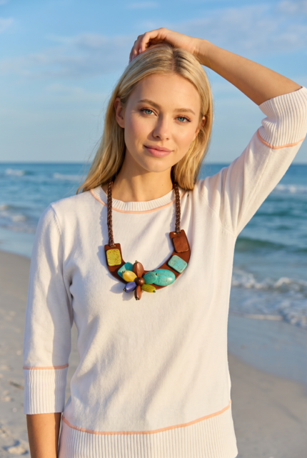 Modella con un collo di perle colorate e una maglietta bianca, in piedi sulla spiaggia con il mare sullo sfondo. La luce del sole illumina la scena, creando un'atmosfera estiva e fresca.