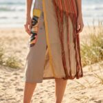 Gonna estiva creativa con dettagli colorati, indossata da una donna sulla spiaggia, in un ambiente soleggiato. La gonna presenta un design unico con strisce e frange, abbinata a sandali.