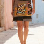 Gonna colorata con frange indossata da una donna in una strada soleggiata, abbinata a una maglietta bianca e sandali. Sfondo di edifici con pareti colorate.