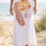 Gonna lunga bianca con dettagli artistici, indossata da una donna su una spiaggia sabbiosa, con il mare sullo sfondo. La gonna presenta un design unico con motivi colorati e tessuti sovrapposti, perfetta per un look estivo elegante e informale.
