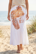 Gonna lunga bianca con dettagli artistici, indossata da una donna su una spiaggia sabbiosa, con il mare sullo sfondo. La gonna presenta un design unico con motivi colorati e tessuti sovrapposti, perfetta per un look estivo elegante e informale.