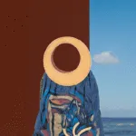 Borsa di design con manico rotondo in pelle, realizzato in tessuto blu con motivi colorati e decorazioni floreali, su sfondo marino e cielo sereno.