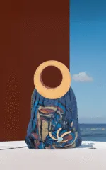 Borsa di design con manico rotondo in pelle, realizzato in tessuto blu con motivi colorati e decorazioni floreali, su sfondo marino e cielo sereno.