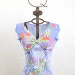 Maglietta blu con stampa floreale su manichino, caratterizzata da un design avvolgente e dettagli colorati. Perfetta per la stagione estiva, con scollatura a V e maniche corte.