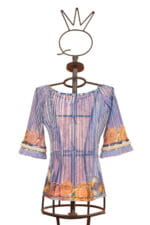 Blusa elegante a maniche lunghe con motivi floreali colorati, presentata su un manichino artistico. Design moderno con dettagli in frange e tonalità di blu e arancione. Ideale per occasioni casual e primaverili.