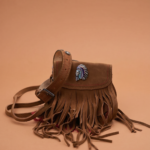 Borsa in pelle scamosciata marrone con frange e decorazione a forma di capo indiano, su sfondo colorato. Ideale per uno stile bohemien e casual.