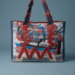 Borsa a spalla colorata con motivi artistici, caratterizzata da manici rossi e dettagli in tessuto blu e bianco, ideale per un look moderno e creativo.
