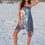Modella sorridente in costume da bagno estivo con un pareo colorato, posata sulla spiaggia bianca al tramonto. Il pareo presenta un design vivace con palme e motivi tropicali, perfetto per una giornata al mare.