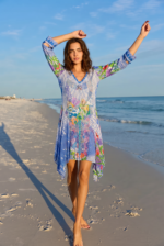 Modella che indossa un vestito estivo colorato con maniche lunghe, mentre cammina lungo una spiaggia al tramonto, con il mare sullo sfondo e il cielo sereno.