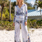 Modella in abbigliamento estivo con stampa floreale, indossa una felpa con zip e pantaloni larghi, in piedi su una spiaggia di sabbia bianca, circondata da palme e vegetazione tropicale.