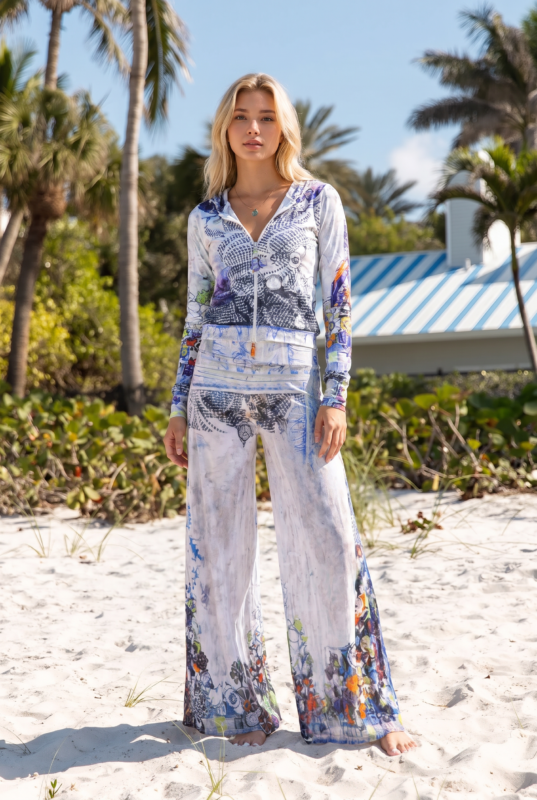 Modella in abbigliamento estivo con stampa floreale, indossa una felpa con zip e pantaloni larghi, in piedi su una spiaggia di sabbia bianca, circondata da palme e vegetazione tropicale.