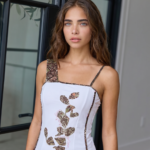 Modella che indossa un top bianco con dettagli floreali e spalline sottili, in un ambiente luminoso con grandi finestre. Il design del top presenta elementi grafici in tonalità naturali, perfetto per un look estivo elegante.