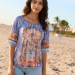 Modella in spiaggia che indossa una blusa colorata con motivi tribali e pantaloni corti in denim, con palme e cielo sereno sullo sfondo.