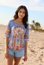 Modella in spiaggia che indossa una blusa colorata con motivi tribali e pantaloni corti in denim, con palme e cielo sereno sullo sfondo.