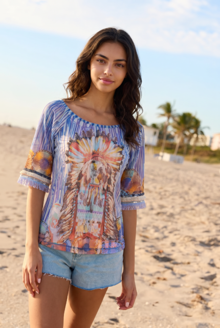 Modella in spiaggia che indossa una blusa colorata con motivi tribali e pantaloni corti in denim, con palme e cielo sereno sullo sfondo.