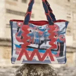 Borsa a tracolla colorata con motivi astratti, appoggiata su una pietra, in un ambiente naturale. Design moderno e creativo, ideale per un look casual e artistico.