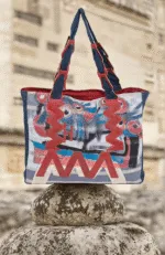 Borsa a tracolla colorata con motivi astratti, appoggiata su una pietra, in un ambiente naturale. Design moderno e creativo, ideale per un look casual e artistico.