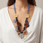 Collana colorata indossata da una donna, composta da perline viola e pendenti decorativi in plastica, tra cui frutti e simboli, su sfondo neutro.