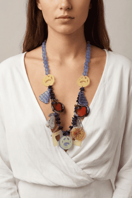 Collana colorata indossata da una donna con maglietta bianca, composta da perline trasparenti blu e pendenti artistici in diverse forme e colori, tra cui frutta e simboli grafici.