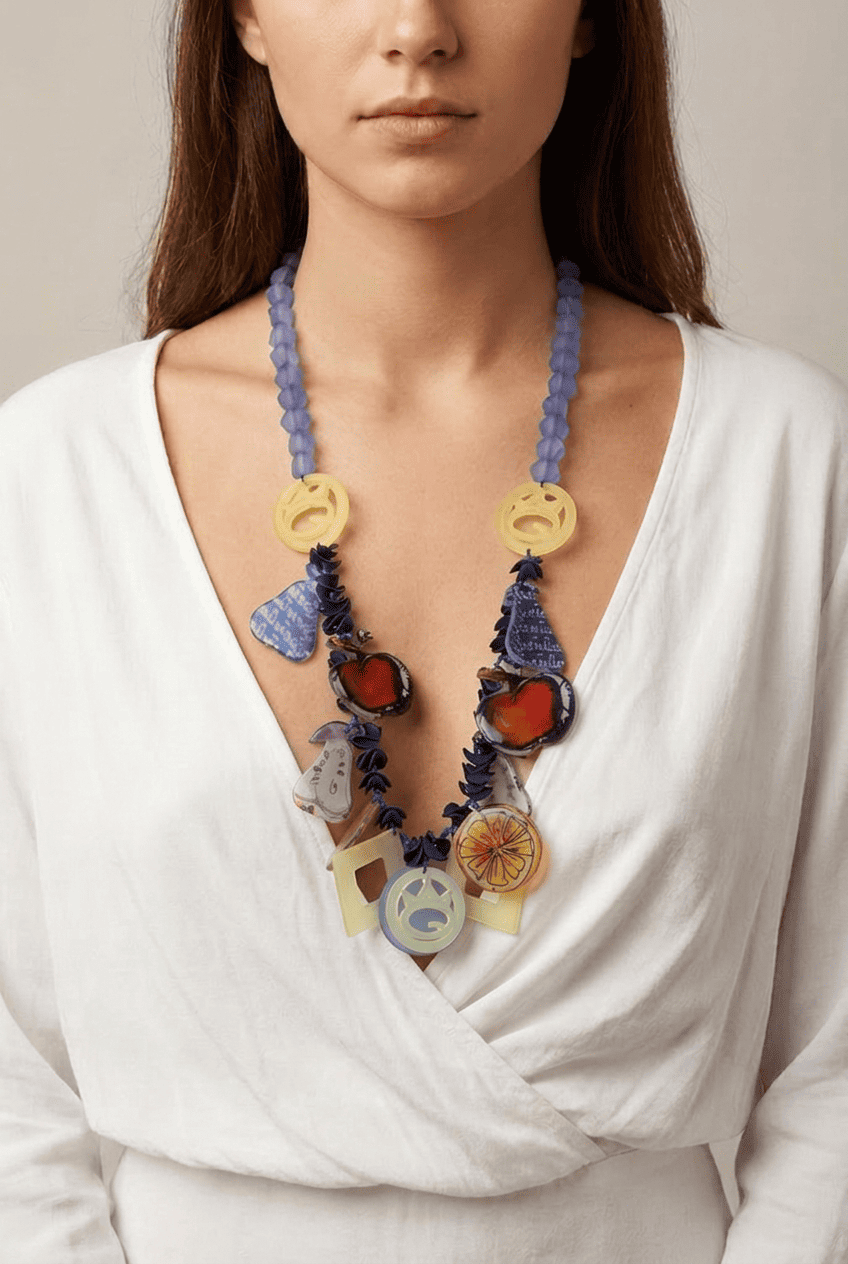 11SSNG902_model Collana colorata indossata da una donna con maglietta bianca, composta da perline trasparenti blu e pendenti artistici in diverse forme e colori, tra cui frutta e simboli grafici.