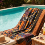 Asciugamano colorato su una sdraio accanto a una piscina, circondato da piante fiorite. Un cappello di paglia e un bicchiere di cocktail sono posizionati su un tavolino in legno.