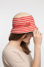 Cappello cloche in cotone a righe rosse e beige indossato da una donna, in primo piano, con le mani che toccano il cappello.