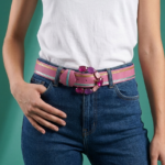 Cintura rosa in vera pelle con fibbia decorata e pietre sintetiche, indossata da una donna in t-shirt bianca e jeans blu, su sfondo verde.