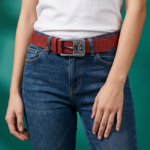 Cintura rossa in vera pelle con fibbia logata indossata da una donna, abbinata a una maglietta bianca e jeans blu, su sfondo verde.