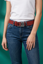 Cintura rossa in vera pelle con fibbia logata indossata da una donna, abbinata a una maglietta bianca e jeans blu, su sfondo verde.