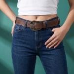 Cintura in vera pelle marrone con borchie e dettagli loggati, indossata da una donna con jeans blu e t-shirt bianca, su sfondo verde.