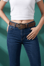 Cintura in vera pelle marrone con borchie e dettagli loggati, indossata da una donna con jeans blu e t-shirt bianca, su sfondo verde.
