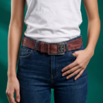 Cintura in vera pelle con maxi fibbia indossata da una donna, abbinata a una t-shirt bianca e jeans blu, su sfondo verde.