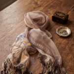 Sciarpa in seta stampata nei toni dei marroni, drappeggiata su un tavolo di legno, con dettagli ricamati e frange decorative, accanto a una scatola nera e un vassoio con monete.