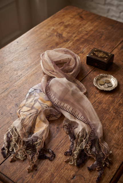 Sciarpa in seta stampata nei toni dei marroni, drappeggiata su un tavolo di legno, con dettagli ricamati e frange decorative, accanto a una scatola nera e un vassoio con monete.