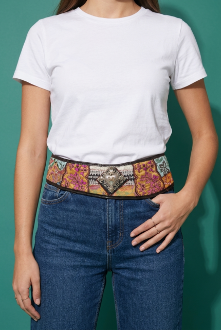 Cintura in tessuto ricamato con dettagli in pelle stampata, indossata da una donna con t-shirt bianca e jeans, su sfondo verde.