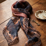 Foulard in crepon di seta con orlo roulotté, caratterizzato da una stampa geometrica in tonalità blu, marrone e rosa, posato su un tavolo di legno accanto a una chiave e un piattino decorativo.