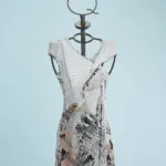 Abito estivo elegante in tessuto leggero, presentato su un manichino con supporto artistico. Il vestito presenta un design innovativo con dettagli floreali e geometrici, ideale per occasioni casual e formali. Sfondo azzurro che mette in risalto il capo.