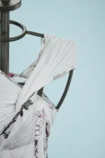 Dettaglio di un abito estivo con stampa floreale, appeso a una gruccia su sfondo blu. Il tessuto bianco presenta drappeggi eleganti e un design moderno, ideale per occasioni informali.