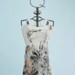 Abito estivo a maniche scoperte con stampe artistiche e colori vivaci, esposto su un manichino con supporto decorativo. Sfondo azzurro chiaro. Ideale per occasioni casual e moda estiva.