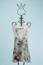 Abito estivo a maniche scoperte con stampe artistiche e colori vivaci, esposto su un manichino con supporto decorativo. Sfondo azzurro chiaro. Ideale per occasioni casual e moda estiva.