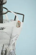 Top bianco con dettagli artistici e decorazione a forma di conchiglia, appeso su un manichino di moda. Sfondo azzurro per un contrasto elegante.