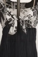 Abito elegante con scollo a V e nodo centrale, caratterizzato da un design grafico in bianco e nero. La parte superiore è in tessuto leggero, mentre la gonna è di un materiale scuro e fluttuante, ideale per occasioni estive.