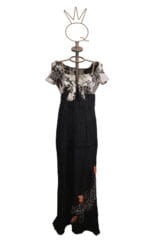 Abito lungo da donna con maniche corte e scollatura a V, caratterizzato da un top in bianco e nero con motivo floreale e una gonna nera con ricami colorati. Esposto su un manichino con una base decorativa.