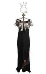 Abito lungo da donna con maniche corte e scollatura a V, caratterizzato da un top in bianco e nero con motivo floreale e una gonna nera con ricami colorati. Esposto su un manichino con una base decorativa.