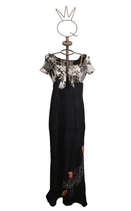 Abito lungo da donna con maniche corte e scollatura a V, caratterizzato da un top in bianco e nero con motivo floreale e una gonna nera con ricami colorati. Esposto su un manichino con una base decorativa.